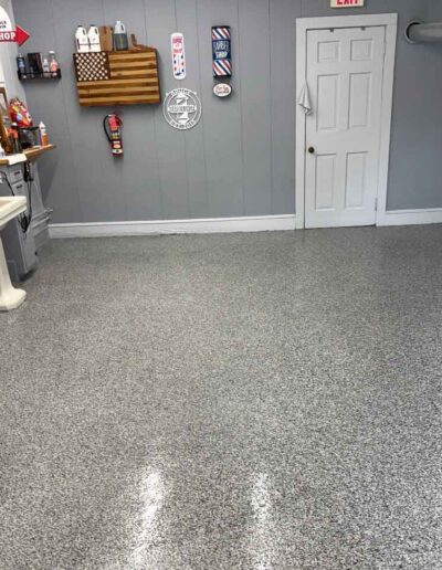 A spacious room featuring a shiny, speckled gray epoxy floor, decorated on one side with barbershop equipment, vintage signs, and white pedestal sinks - www.shoreconcretecoating.com