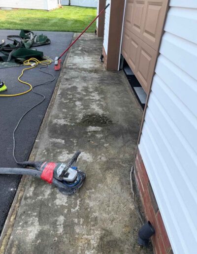 An angle grinder attached to a vacuum hose rests on a worn concrete walkway outside a residential garage - www.shoreconcretecoating.com