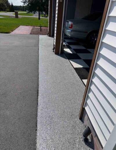 A textured gray speckled coating lines the apron between a dark asphalt driveway and an open garage - www.shoreconcretecoating.com